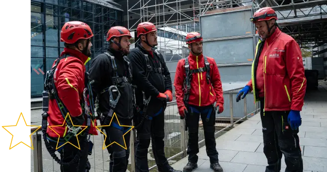 Fünf Gerüstbau-Mitarbeiter der Firma MACK in Arbeitskleidung mit Helm und Sicherheitsgurt im Team-Gespräch mit Projektleiter auf der Baustelle.