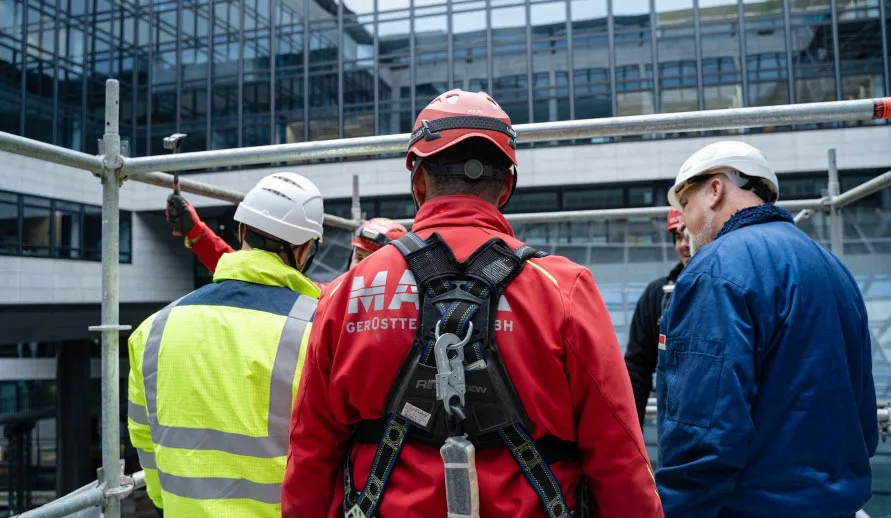 Fünf Personen stehen auf einem Gerüst. Drei davon sind MACK-Kollegen, während drei Personen mit dem Rücken zur Kamera zu sehen sind.