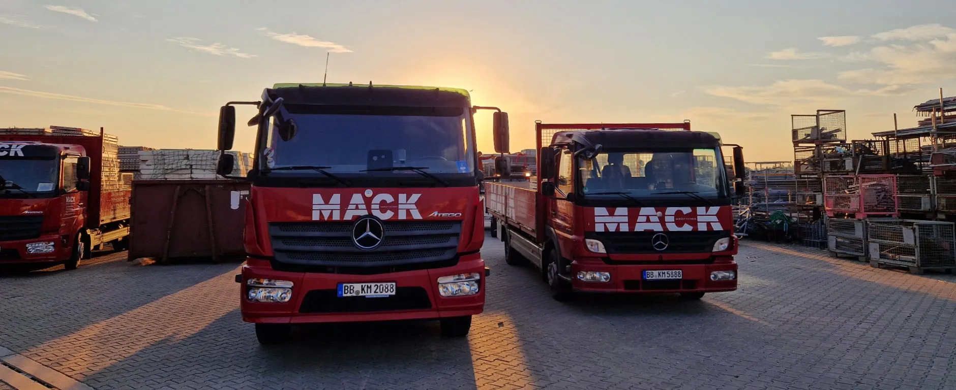 Zwei rote LKW-Pritschen mit MACK-Logo auf dem Firmengelände, im Hintergrund weitere Fahrzeuge und das Gerüstbaulager.