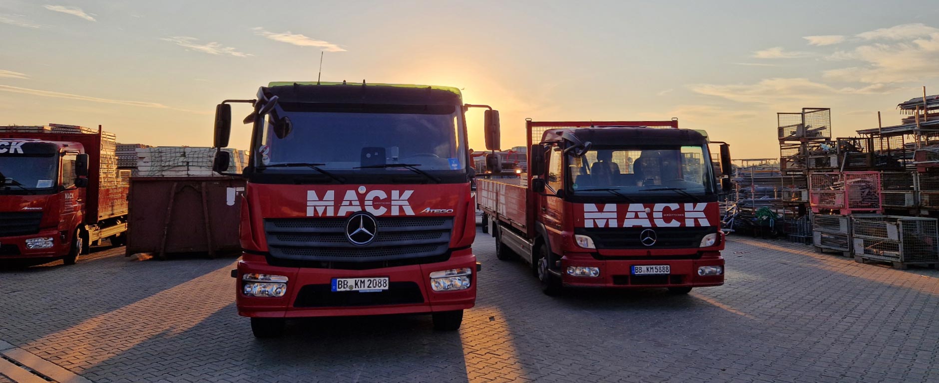 Zwei rote LKW-Pritschen mit MACK-Logo auf dem Firmengelände, im Hintergrund weitere Fahrzeuge und das Gerüstbaulager.