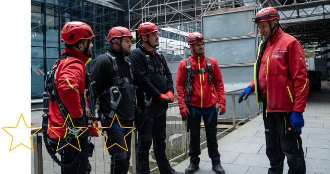 Fünf Gerüstbau-Mitarbeiter der Firma MACK in Arbeitskleidung mit Helm und Sicherheitsgurt im Team-Gespräch mit Projektleiter auf der Baustelle.