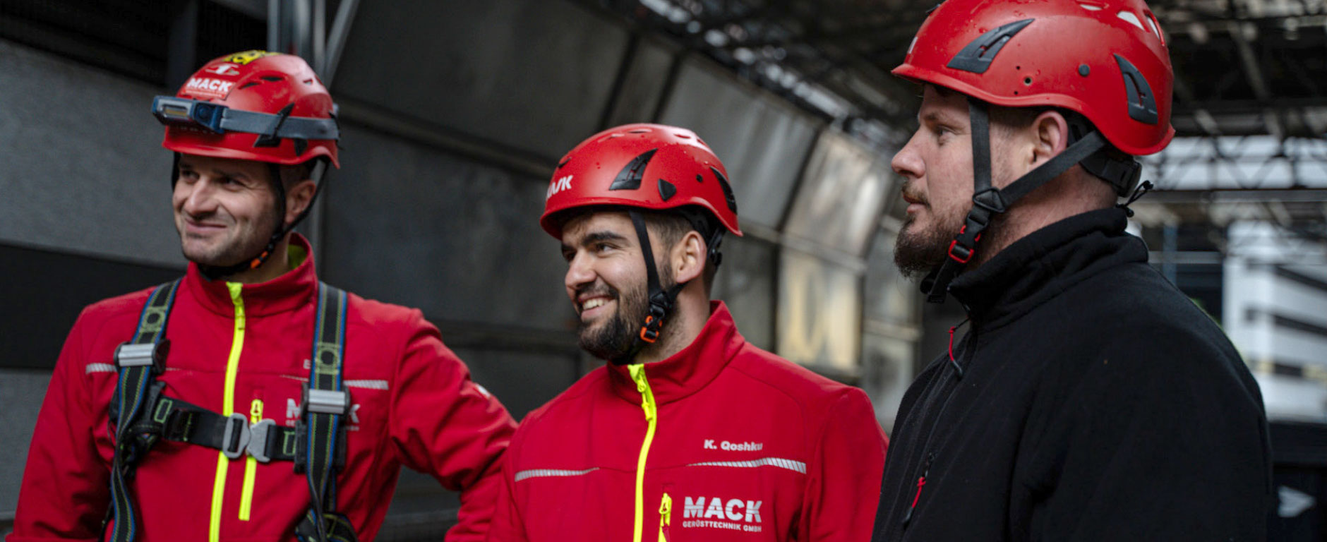 Drei Gerüstbau-Mitarbeiter der Firma MACK in Arbeitskleidung im Team-Gespräch auf der Baustelle.